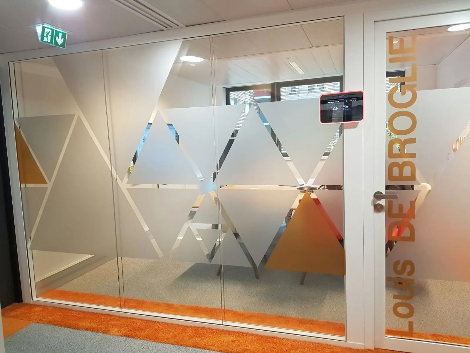 Salle de réunion vitrée habillée de triangles de couleur et dépolis, avec son nom en grandes lettres adhésives sur la cloison